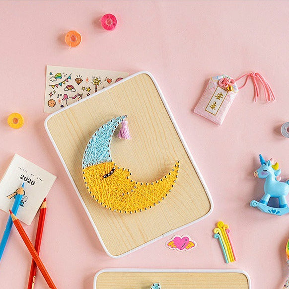 A children's craft board featuring a yellow crescent moon and blue stars made of string, surrounded by colorful pencils, stickers, a small unicorn toy, and decorative items on a soft pink background.